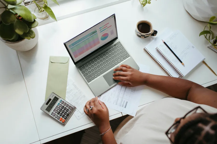 Osoba siedząca przy biurku pracuje na laptopie, obok niej leży kalkulator, notatnik i filiżanka kawy. Na ekranie laptopa widoczne są wykresy i tabele finansowe.