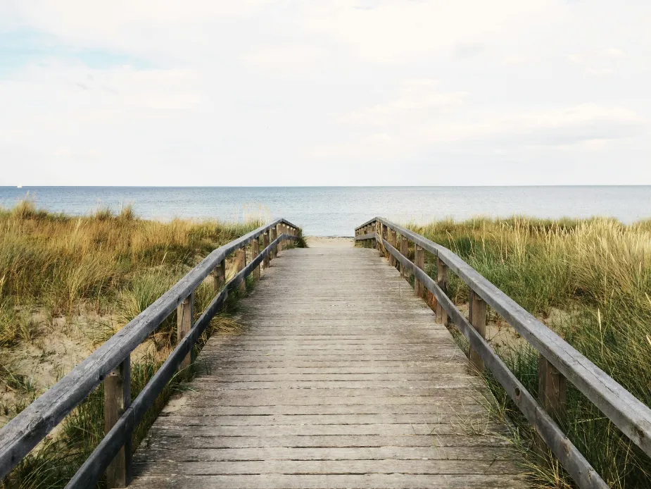 Drewniana ścieżka prowadząca na spokojną plażę Tekst alternatywny: Drewniana ścieżka prowadząca przez wysoką trawę do spokojnej, rozległej plaży, z widokiem na ocean pod częściowo zachmurzonym niebem.