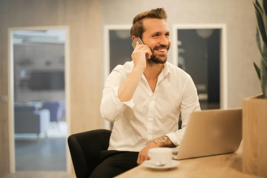 Uśmiechnięty mężczyzna w białej koszuli rozmawia przez telefon, siedząc przy biurku z laptopem. W tle widać wnętrze biura.