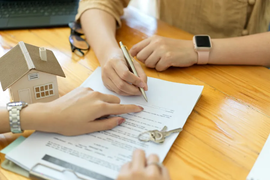 Dwie osoby podpisujące dokumenty z małym modelem domu i kluczami na drewnianym stole
