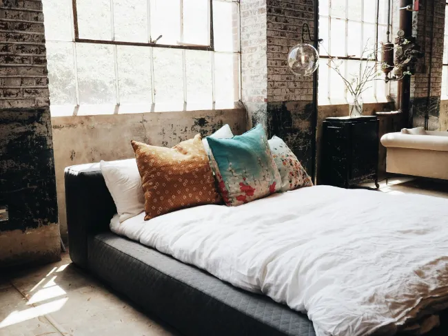 Przestronna sypialnia w stylu industrialnym z dużymi oknami, eksponowaną cegłą, dużym łóżkiem i kolorowymi poduszkami.
