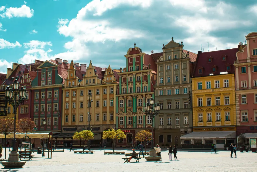 Kolorowe kamienice na rynku w Wrocławiu. Plac wypełniony jest przechodniami, a historyczne budynki w pastelowych kolorach tworzą malowniczą atmosferę. Na pierwszym planie widoczne są latarnie i ławki.
