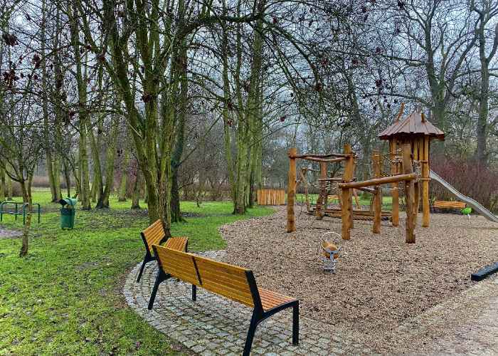 Plac zabaw na osiedlu Bieńkowice we Wrocławiu. Drewniana zjeżdżalnia dla dzieci, otoczona ławkami. Plac zabaw w centrum parku.