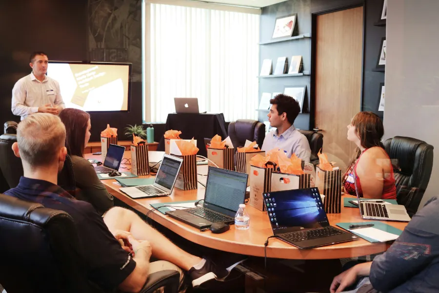 Grupa ludzi uczestnicząca w prezentacji biznesowej, siedząca wokół stołu konferencyjnego z laptopami.