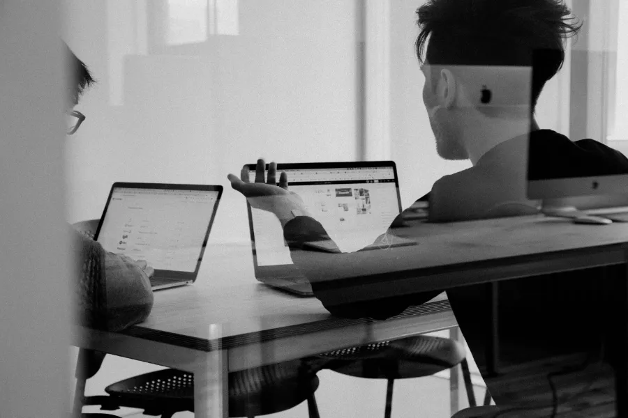 Sylwetka osoby pracującej na dwóch laptopach jednocześnie, siedzącej przy drewnianym stole w biurowym otoczeniu