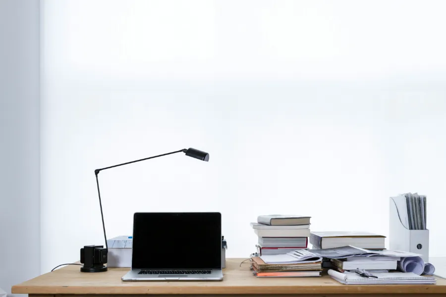 Minimalistyczne miejsce pracy z zamkniętym laptopem, lampką biurkową, stosami książek i dokumentów na drewnianym biurku
