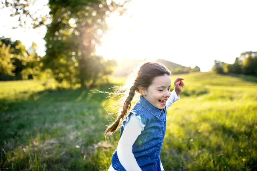 Dziewczynka z warkoczem, ubrana w dżinsową kamizelkę, biega i śmieje się na zielonej łące w promieniach słońca.