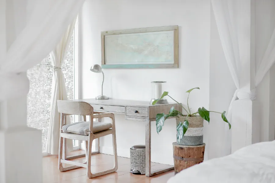  Elegancko urządzone biuro domowe z białymi ścianami i jasnymi zasłonami. Na drewnianym biurku stoi lampa, roślina doniczkowa i dekoracyjna misa.