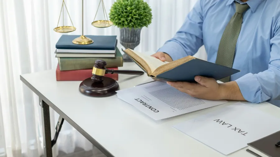 Prawnik przeglądający księgę prawną przy biurku z młotkiem sędziowskim i wagą.