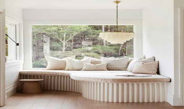 Biała kanapa w niecodziennym kształcie, za która znajduje się duże okno i lasek. 