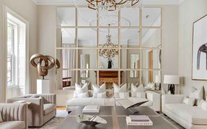 Przestronny pokój, w którym znajdują się białe sofy i białe poduszki, przednia ściana jest wypełniona lustrami w których odbija się żyrandol. 