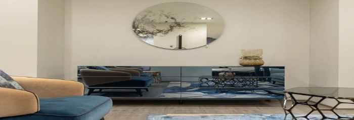 Pod ścianą stoi lustrzana komoda, w której odbija się fotel i nowoczesny stolik. Nad komoda wisi okrągłe lustro. 