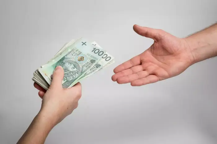 Dłoń jednej osoby przekazuje plik banknotów stuzłotowych drugiej osobie z otwartą, wyciągniętą dłonią