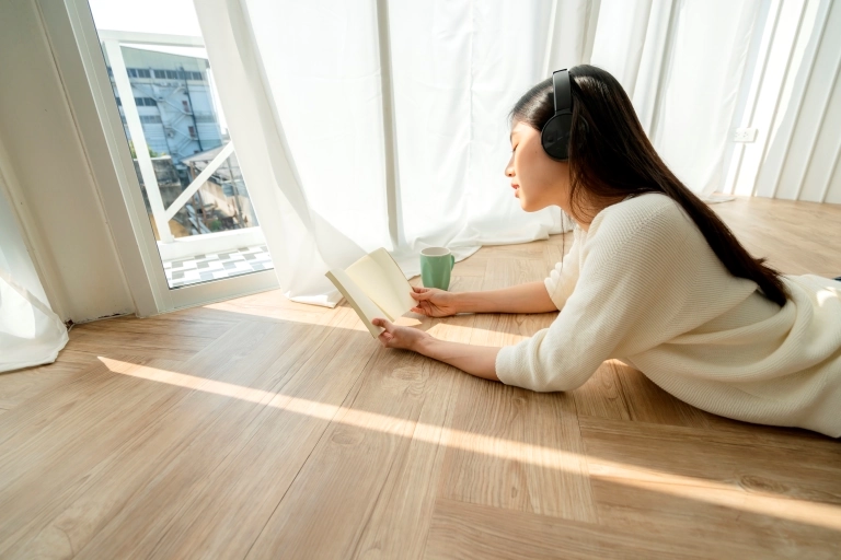 Kobieta leży z książką i zimnym napojem na podłodze przy zasłoniętych oknach