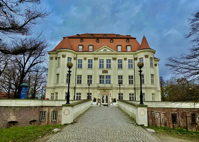 Widok pięknego pałacu znajdującego się w Leśnicy. Kamienna kładka prowadzi do wyremontowanego budynku o jasnej fasadzie i licznych okach, po prawej i lewej dwie wieżyczki.