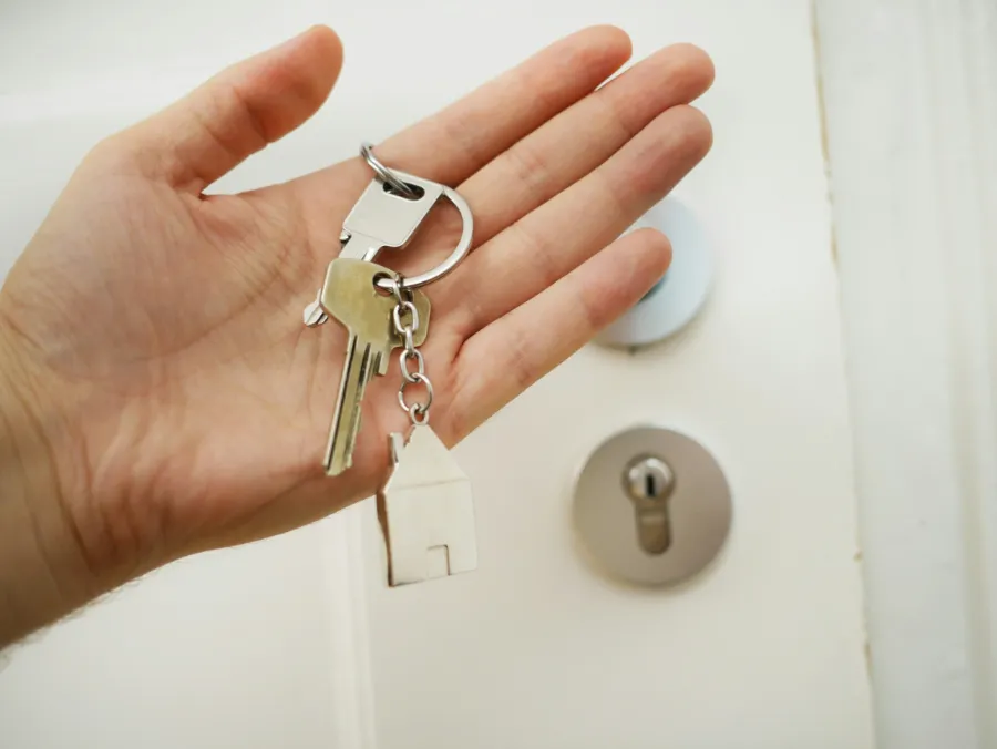 Ręka trzymająca klucze z brelokiem w kształcie domu, gotowe do przekazania, przy białych drzwiach z zamkiem.
