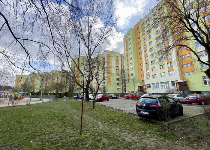 Osiedle mieszkaniowe zabudowane wielkimi, żółtymi blokami na osiedlu Nowy Dwór we Wrocławiu. Przed blokami są miejsca parkingowe oraz samochody.
