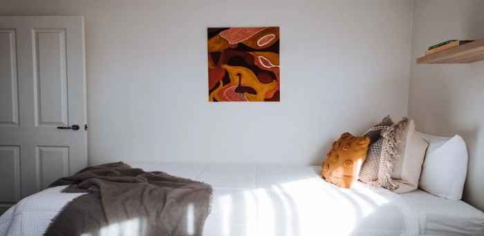 Łóżko, na którym leży lekko pomięty koc i kilka poduszek, nad nim wisi brązowy obraz, do pomieszczania prowadza otwarte białe drzwi. 