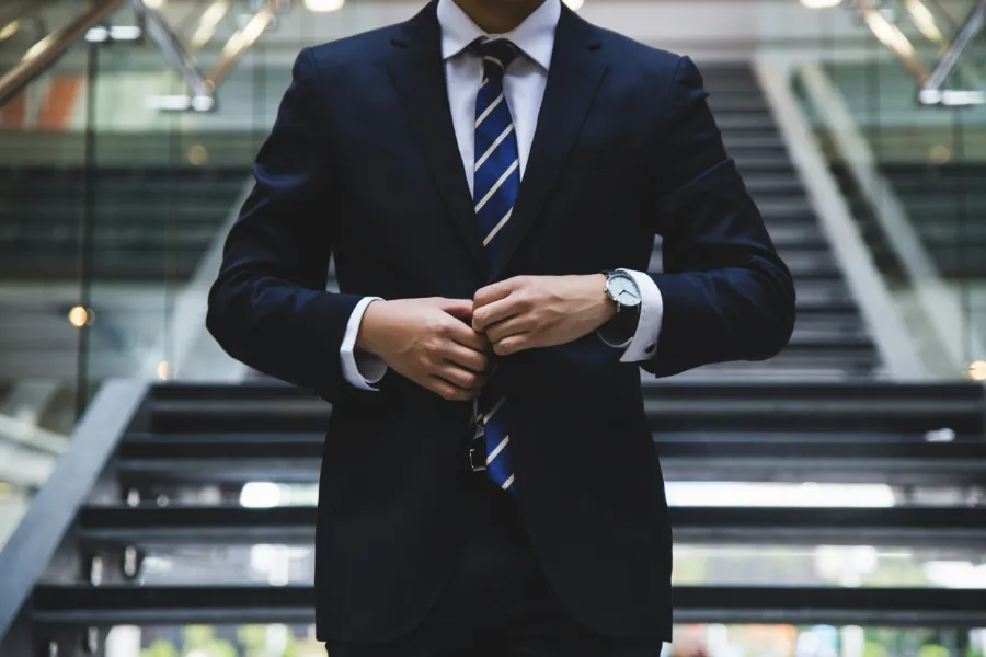 Mężczyzna w eleganckim granatowym garniturze i niebieskim krawacie zapinający marynarkę, stojący na tle schodów ruchomych w nowoczesnym biurowcu.