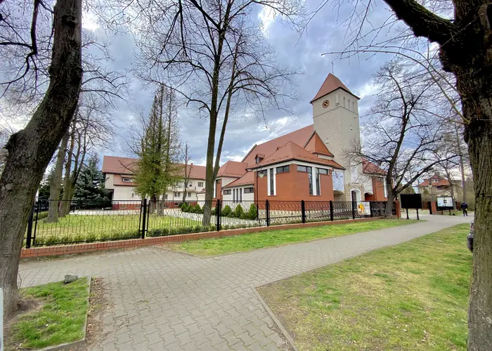 Kościół z czerwonej i jasnej cegły z czerwonymi dachówkami. Przed kościołem chodnik otoczony trawą i drzewami