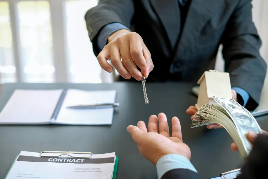 Ręka w czarnym garniturze przekazuje klucze do domu nad stołem z pieniędzmi i dokumentami