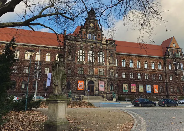 Muzeum narodowe we Wrocławiu, porośnięte bluszczem, z ceglaną neorenesansową fasadą, oraz plakatami przedstawiającymi trwające wystawy. 