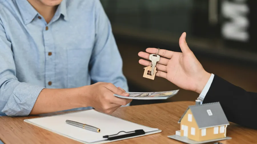 Osoba przekazuje banknoty i odbiera klucze do nieruchomości z rąk innego człowieka przy stole, na którym znajduje się model domu