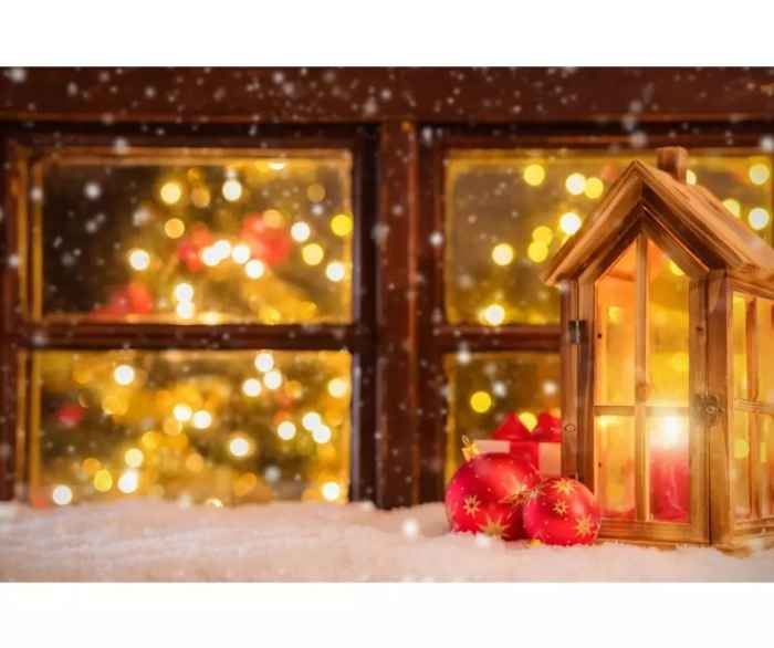 Drewniany lampion i czerwone bombki ze złotymi gwiazdkami leżą na śniegu, dalej jest drewniane okno, zza którego widać rozświetlone choinki. 