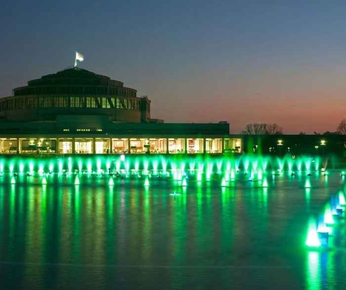Zdjęcie podświetlonej Hali Stulecia porą wieczorową. Na pierwszym planie fontanna podświetlona w kolorach zieleni. 