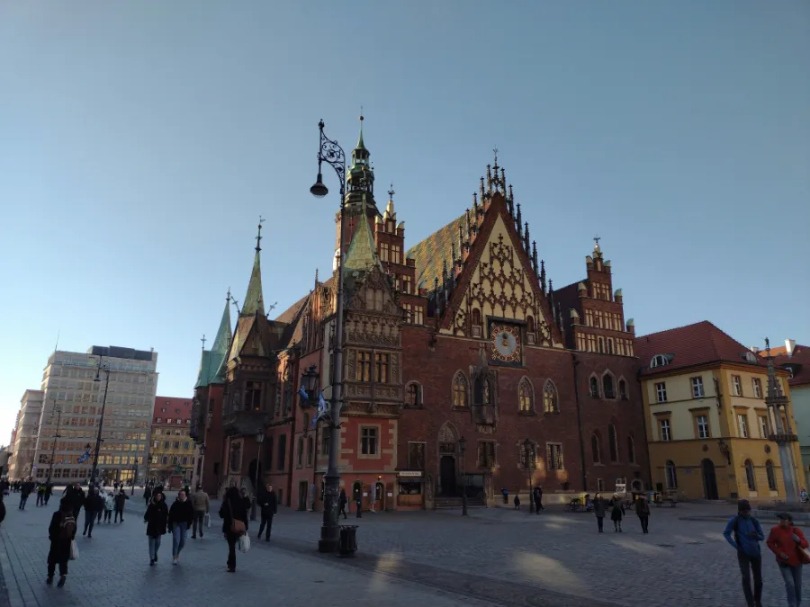 Gotycki Ratusz Wrocławski z czerwono-brunatnymi elewacjami i wysmukłymi wieżami, otoczony przez przechodniów i zabytkowe budynki na Placu Rynku, w słoneczny dzień.